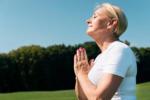 A woman in a white shirt stands outdoors, hands clasped in prayer position, facing the sky with a serene expression against a green background.