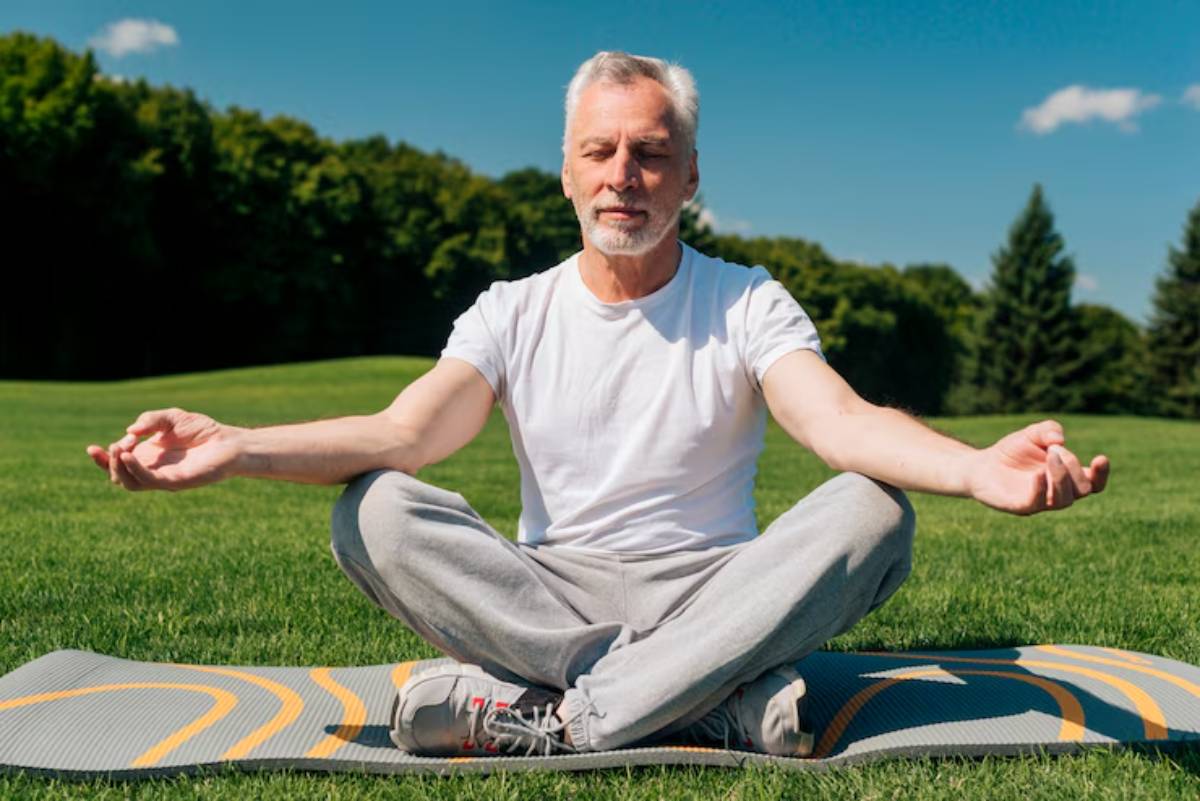 An older man sits cross-legged on a yoga mat in a lush green park, meditating peacefully under a clear blue sky.
