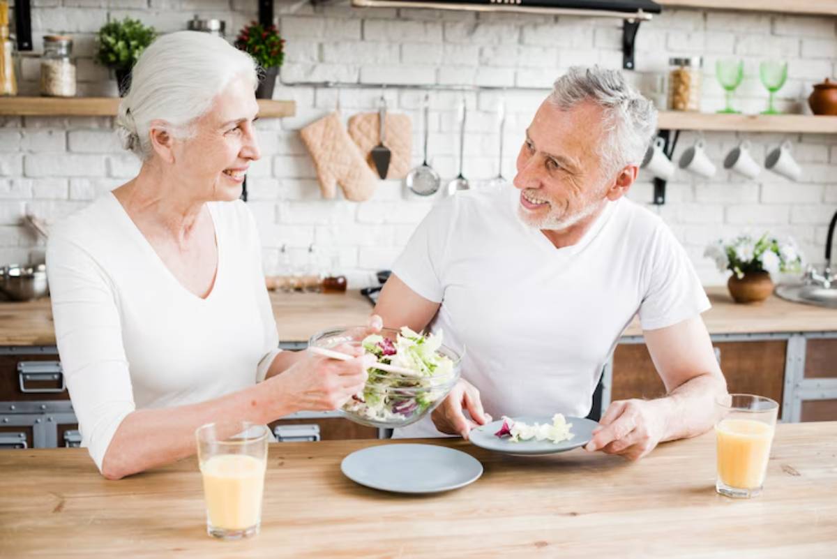 An elderly woman serves a fresh salad to a man at a wooden kitchen table, with glasses of juice placed nearby.