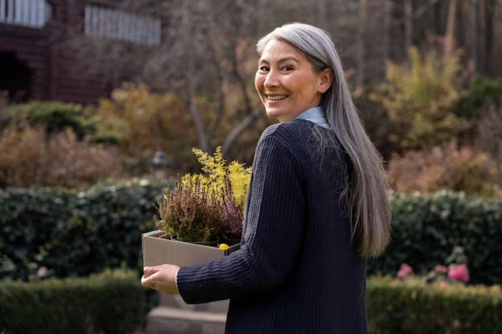 A person wearing a dark sweater holds a box of colorful plants, surrounded by an autumn garden with softly blurred foliage.