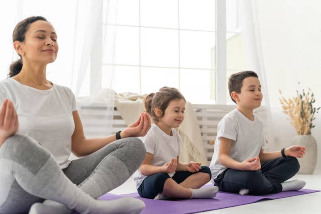 A woman and two children sit cross-legged on a purple mat, practicing meditation in a bright, airy room with large windows.
