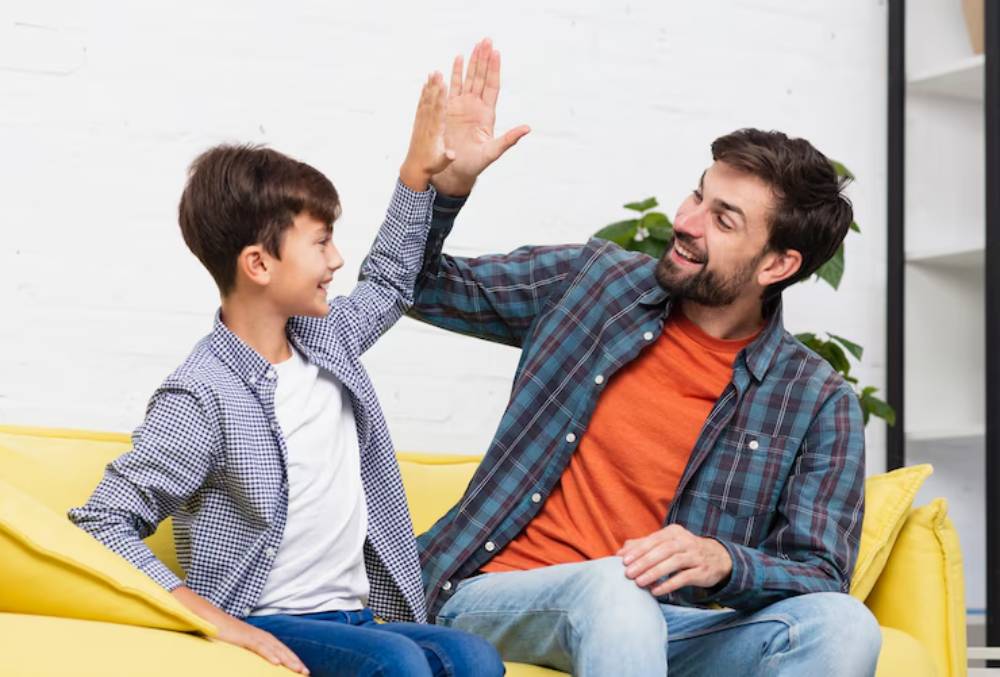 A father and son sit on a yellow couch, engaged in a playful high-five, surrounded by a cozy, modern living space.