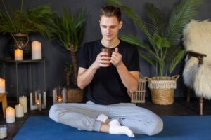 A person sits cross-legged on a yoga mat, holding a cup, surrounded by candles and lush green plants in a tranquil room setting.