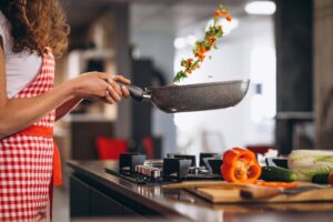 A person cooking in a checkered apron tosses colorful vegetables in a frying pan in a modern kitchen. Fresh ingredients are on the counter.