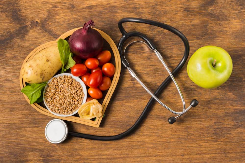 A heart-shaped arrangement of healthy foods including vegetables, grains, and an apple, accompanied by a stethoscope on a wooden surface.