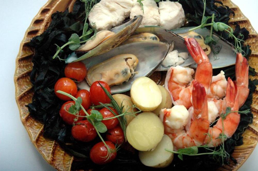 A platter of seafood featuring shrimp, mussels, and fish, accompanied by cherry tomatoes and baby potatoes, garnished with herbs.