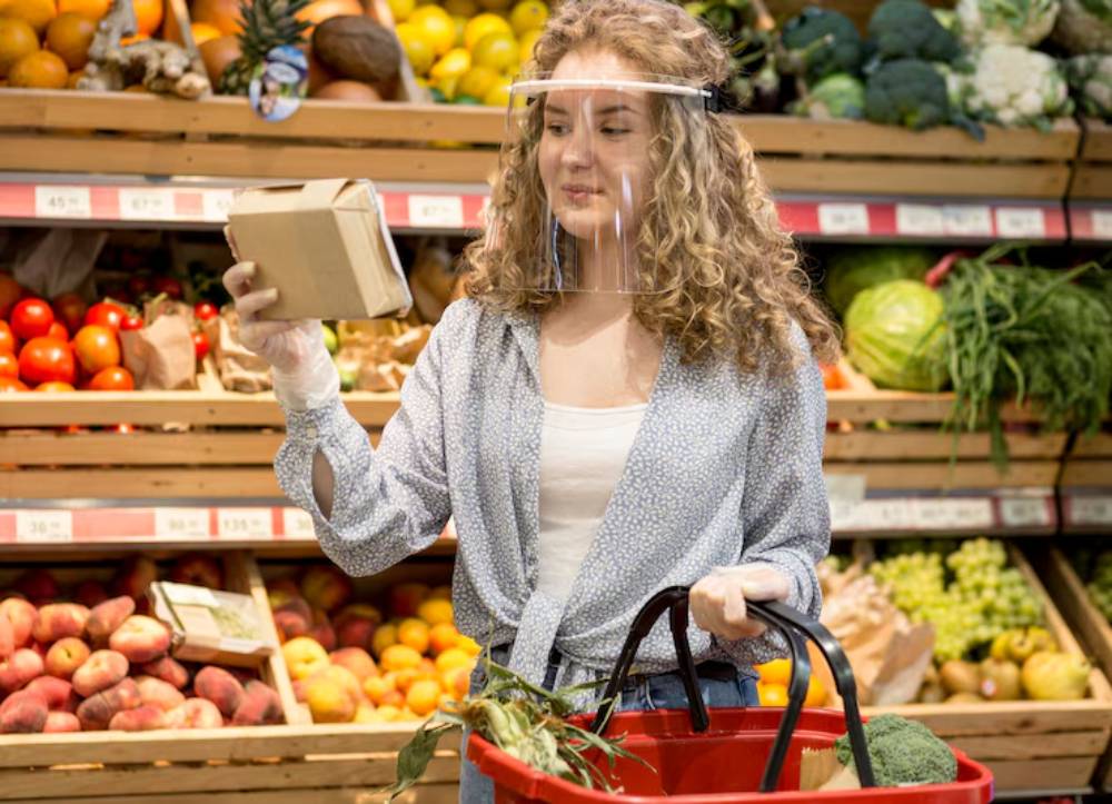 A woman shopping in a grocery store holds a small package while inspecting fresh produce and fruits in her red shopping basket.