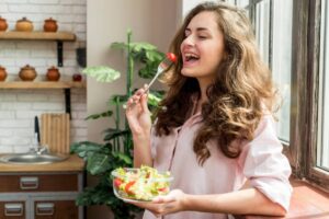 A woman holds a fork with a cherry tomato, standing by a window, while enjoying a bowl of fresh salad in a cozy kitchen.