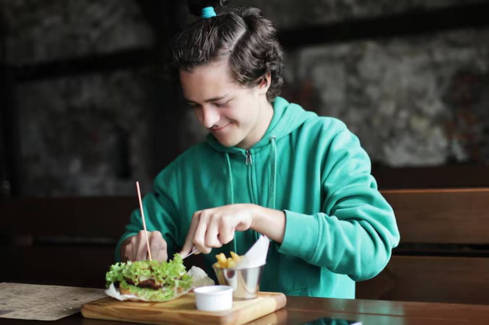 A person in a green hoodie is enjoying a meal of a fresh burger with lettuce and fries, seated at a rustic wooden table.