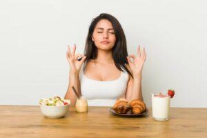 A woman doing mindfulness at table, surrounded by a bowl of salad, a dessert, croissants, and a glass of strawberry milkshake.