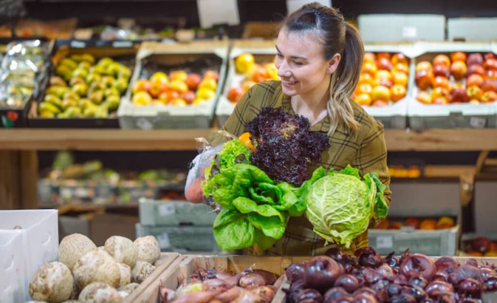 A person holds various fresh greens, including lettuce and cabbage, in a vibrant market filled with colorful fruits and vegetables.