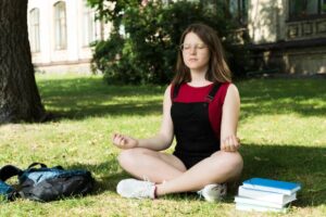 A girl student sits cross-legged on grass in a park, meditating with books stacked beside them and a backpack nearby.