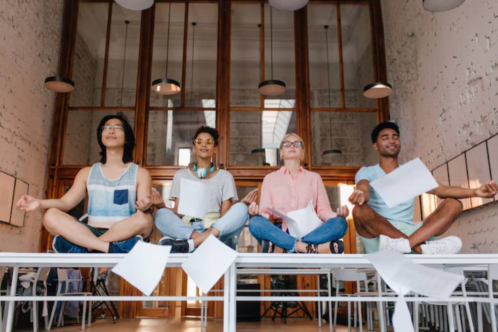 Four individuals sit cross-legged on a table in a modern, airy space, practicing meditation with scattered sheets of paper around them.