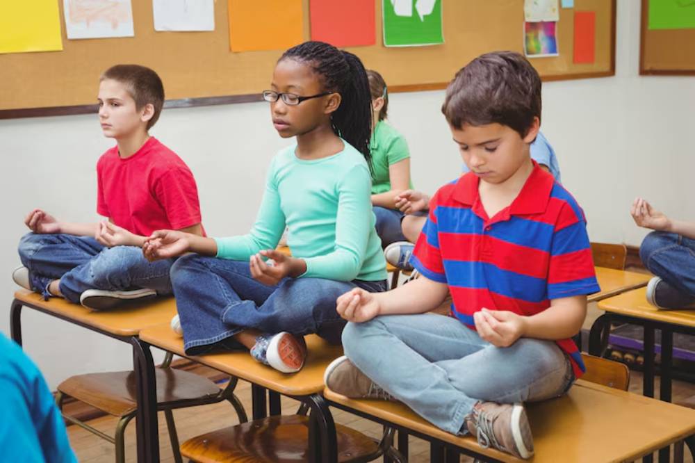 Children sitting cross-legged on desks, practicing mindfulness in a classroom with colorful artwork on the walls.