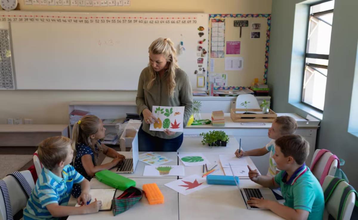 A teacher stands with leaf illustrations, while students engage with laptops and artwork at a classroom table. Bright and organized setting.