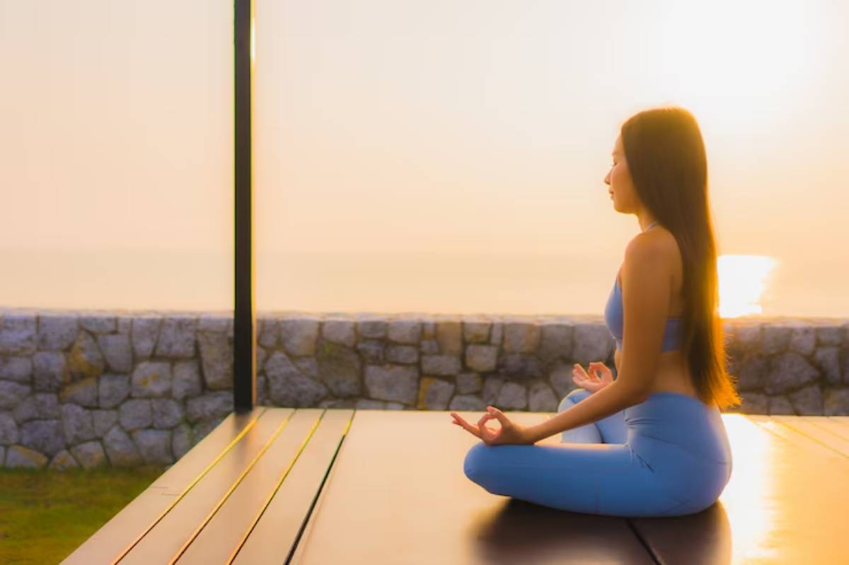 A person sits in a meditative pose at sunrise, overlooking a serene coastline and a stone wall in a peaceful outdoor setting.