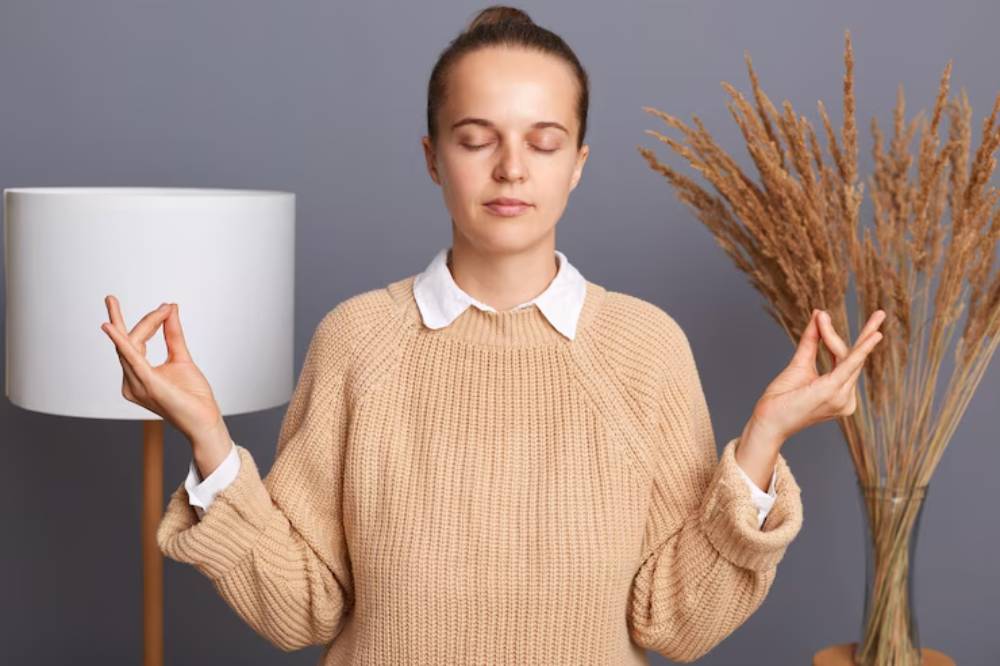 A person in a beige sweater and white collar meditates with hands in a yoga pose, surrounded by a lamp and dried plants.