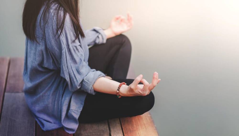 A woman sits cross-legged on a wooden platform, meditating with open hands, wearing a relaxed gray shirt and a beaded bracelet.
