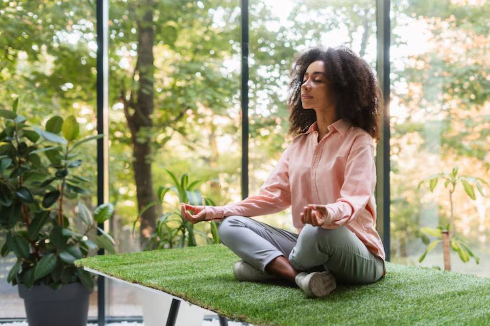 A woman meditates on a grass-covered table surrounded by lush greenery and large windows, creating a serene atmosphere.