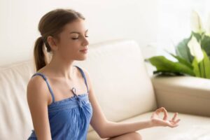 A person sitting cross-legged on a sofa, practicing meditation, wearing a blue tank top, surrounded by indoor plants.