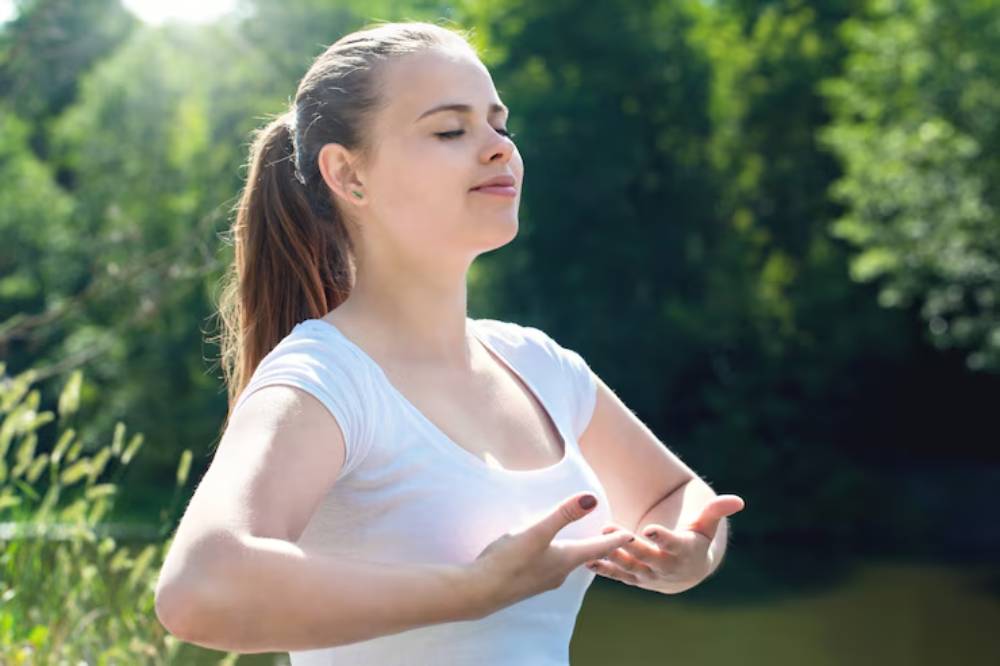 A person in a white shirt stands by a serene body of water, surrounded by lush greenery, with hands raised in a meditative pose.