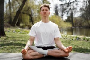 A person sits cross-legged on a yoga mat outdoors, surrounded by greenery and a tranquil water backdrop, practicing meditation.