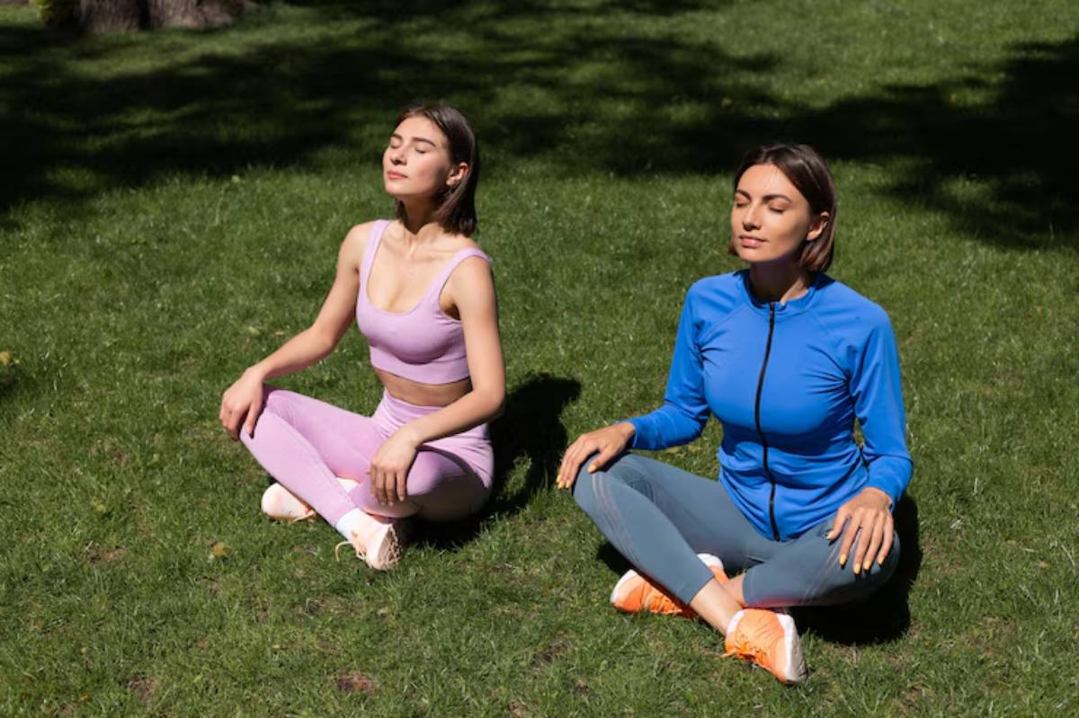 Two women sit cross-legged on green grass, wearing activewear in shades of pink and blue, enjoying a sunny outdoor moment.