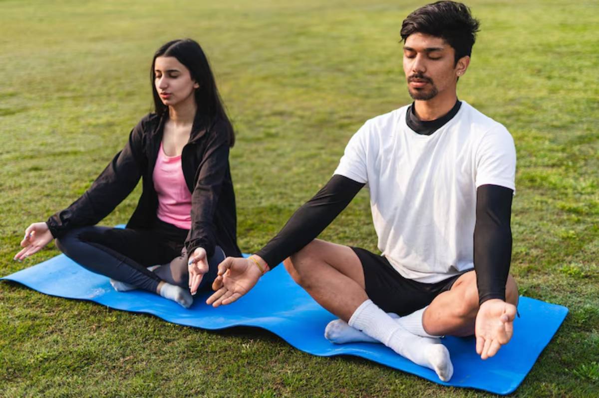A woman and a man practice yoga meditation on a blue mat in a grassy outdoor setting, wearing comfortable athletic clothing.