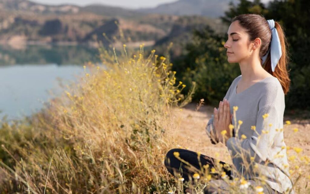 A woman sits in meditation by a serene lake, surrounded by tall grasses and wildflowers, with mountains in the background.