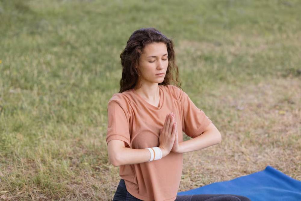 A person in a brown t-shirt practices yoga outdoors, seated on a blue mat with hands in a prayer position, surrounded by grass.