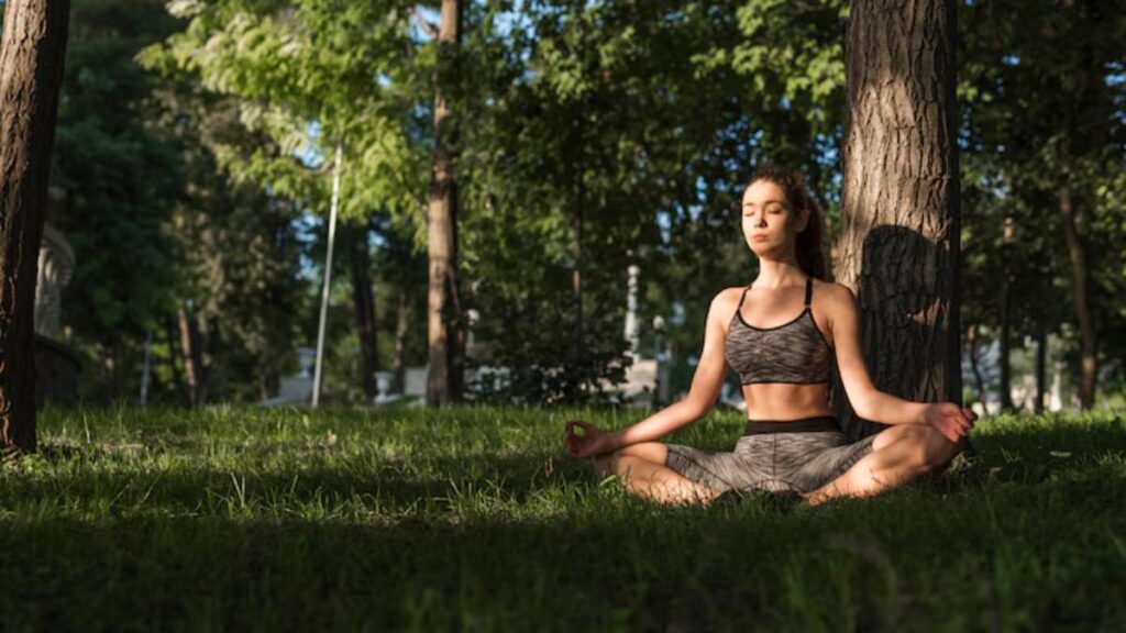 A person sits cross-legged on green grass, practicing yoga in a serene outdoor setting surrounded by tall trees and soft sunlight.