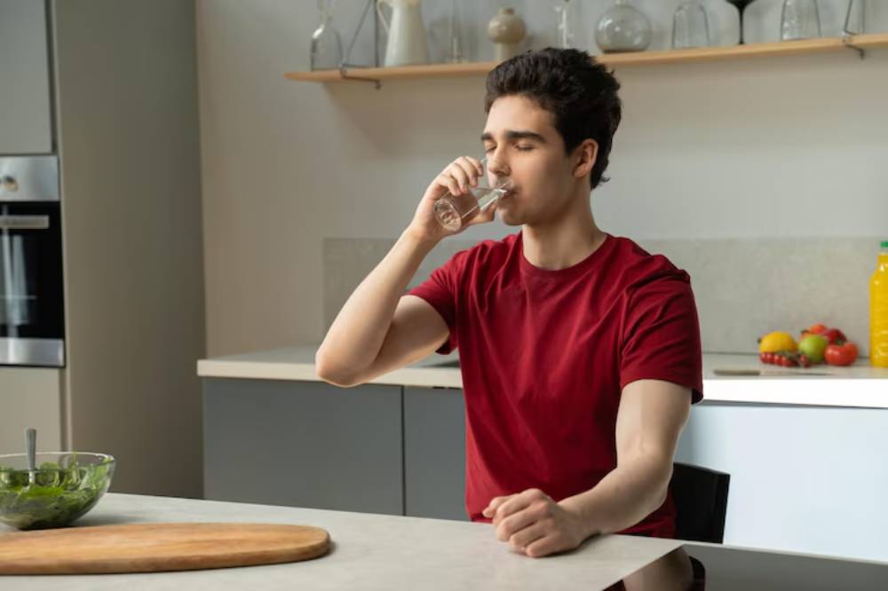 A young man in a red shirt sits at a modern kitchen table, holding a glass of water, with salad and fruits nearby.