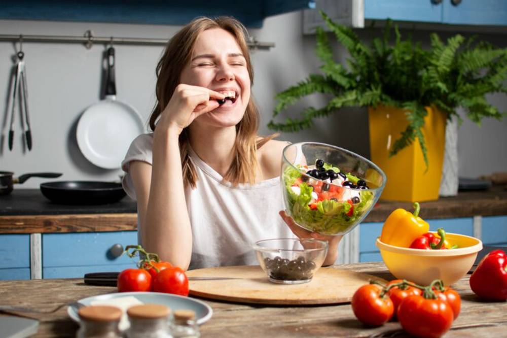 A woman holds a large bowl of colorful salad in a bright kitchen, surrounded by fresh vegetables and cooking utensils.