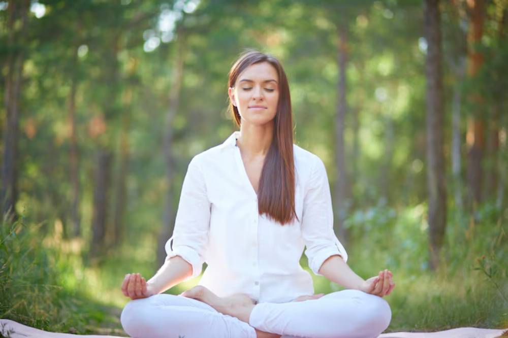 A woman sits cross-legged on a mat in a serene forest, practicing meditation, surrounded by lush greenery and soft sunlight.