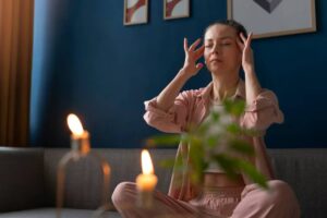 A person sitting cross-legged on a couch in a cozy room, surrounded by candles and greenery, creating a calm atmosphere.