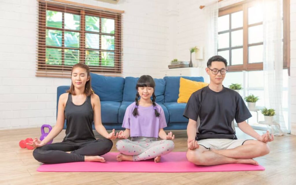 A woman, girl, and man meditate on yoga mats in a bright room with a blue couch and plants, promoting relaxation and mindfulness.