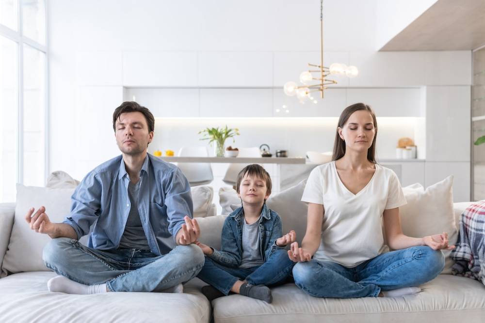 A family of three sits cross-legged on a sofa, practicing meditation in a modern, bright living room with a stylish chandelier.