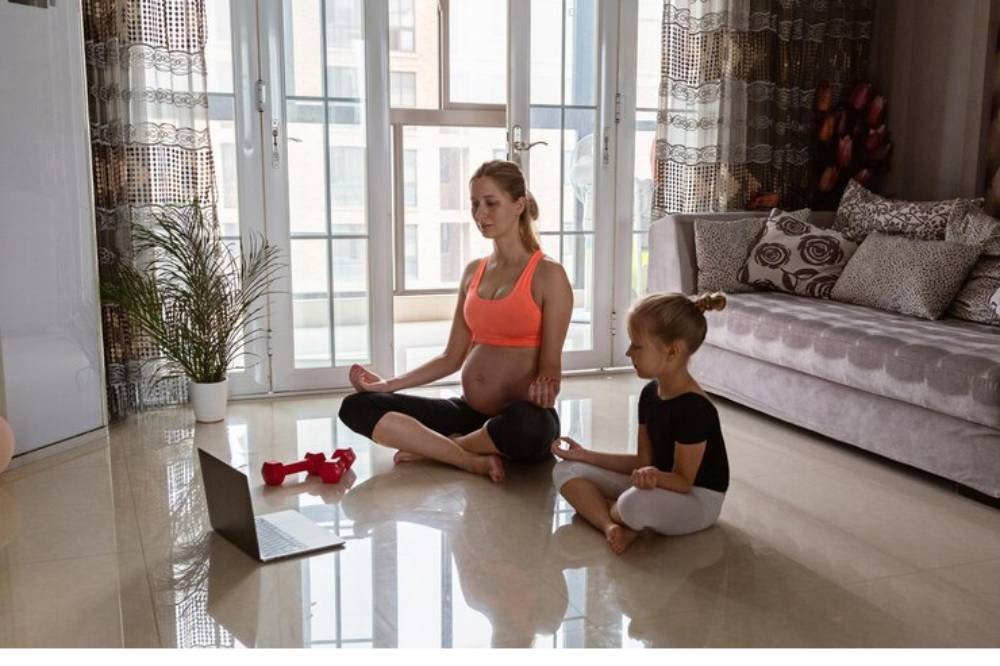 A pregnant woman and a young girl sit cross-legged on a polished floor, practicing mindfulness together in a bright, cozy living room.