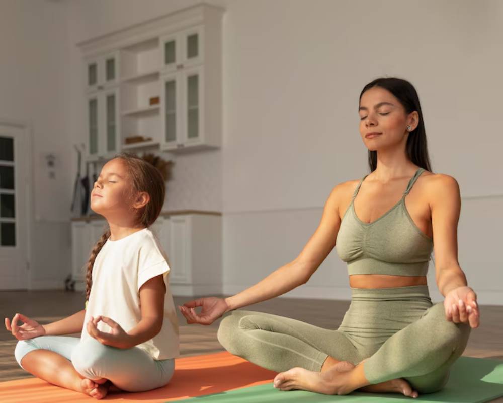 A woman and girl practice yoga on mats in a bright, minimalistic room, engaged in meditation with hands resting on their knees.
