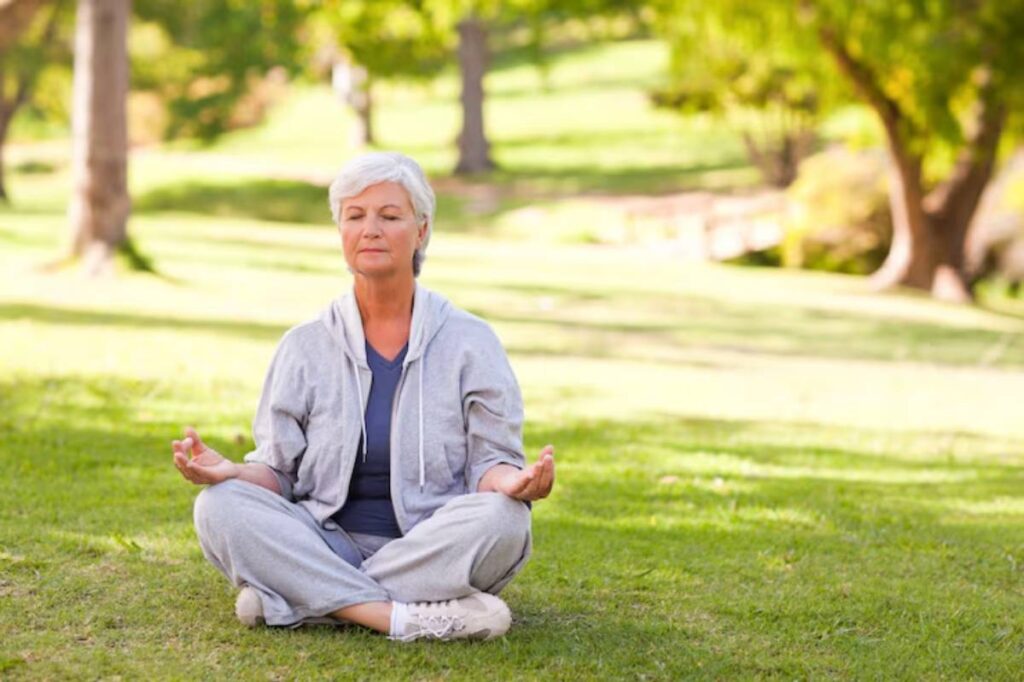 A person sits cross-legged on green grass in a serene park, dressed in a gray tracksuit, practicing meditation.