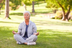 A person sits cross-legged on green grass in a serene park, dressed in a gray tracksuit, practicing meditation.