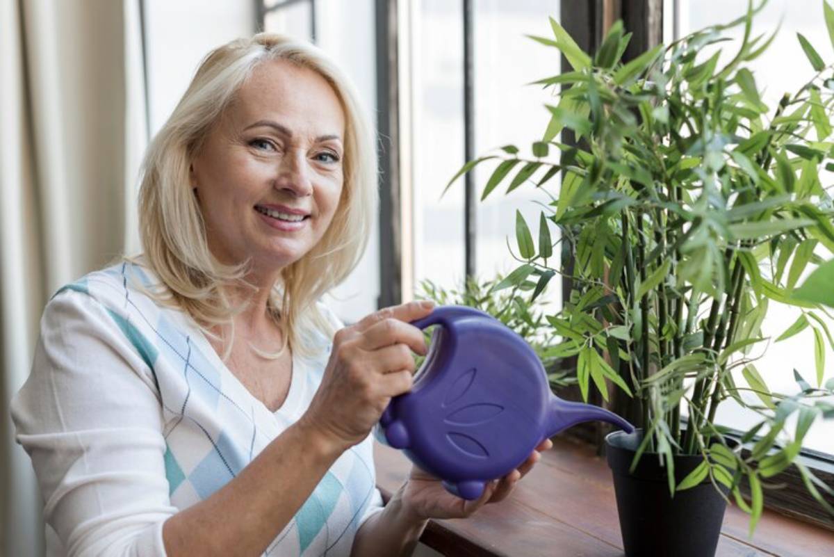 A woman with long blonde hair holds a purple watering can, tending to a green plant by a sunlit window.