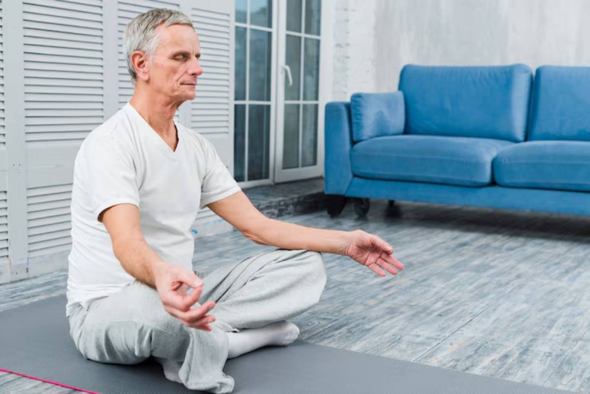 A senior man sits cross-legged on a yoga mat in a cozy room, meditating peacefully in front of a blue couch and bright windows.