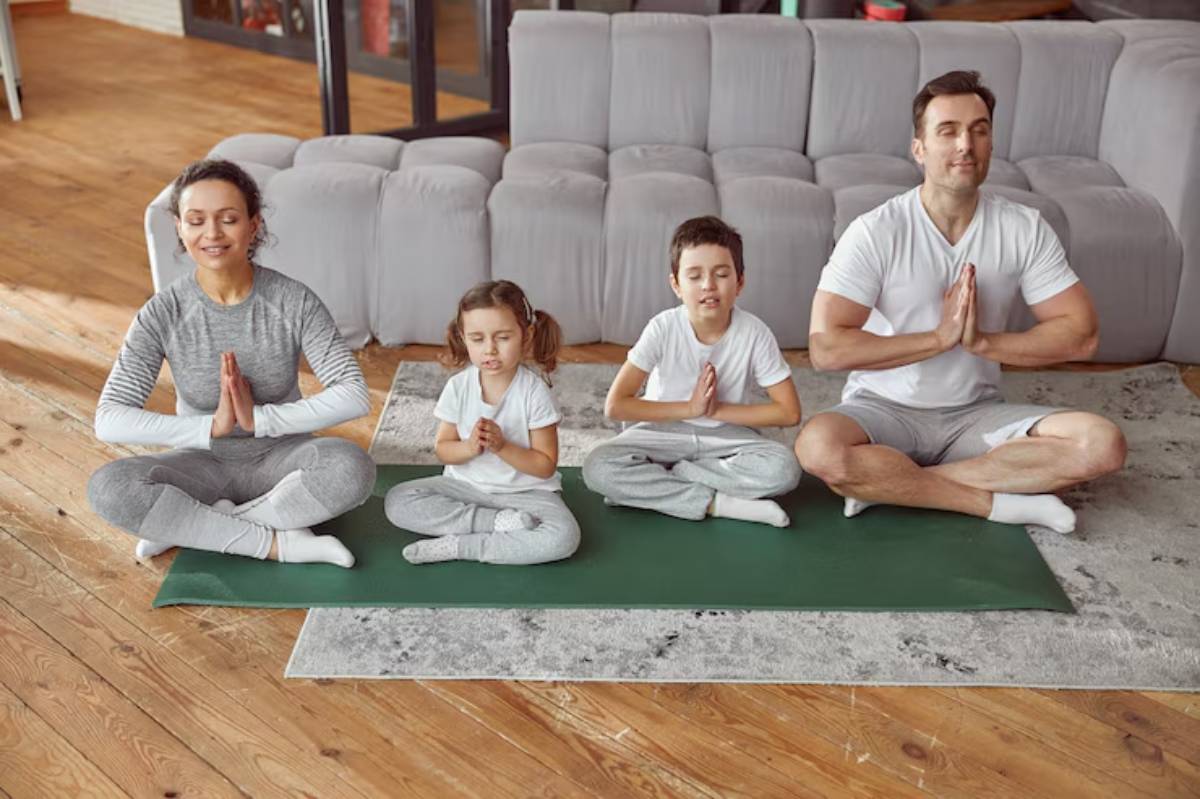 A family of four practices yoga together on green mats in a cozy living room with wooden floors and a gray couch in the background.