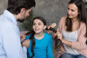 A woman gently styles a girl's hair while a man assists, creating a warm and caring family moment in a cozy setting.
