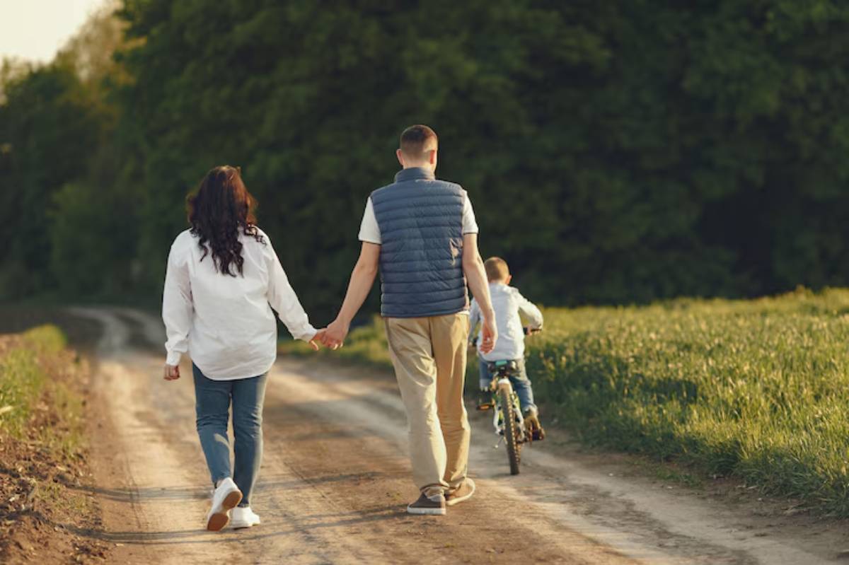 A couple walks hand-in-hand along a dirt path, while their child rides a bike nearby, surrounded by greenery.