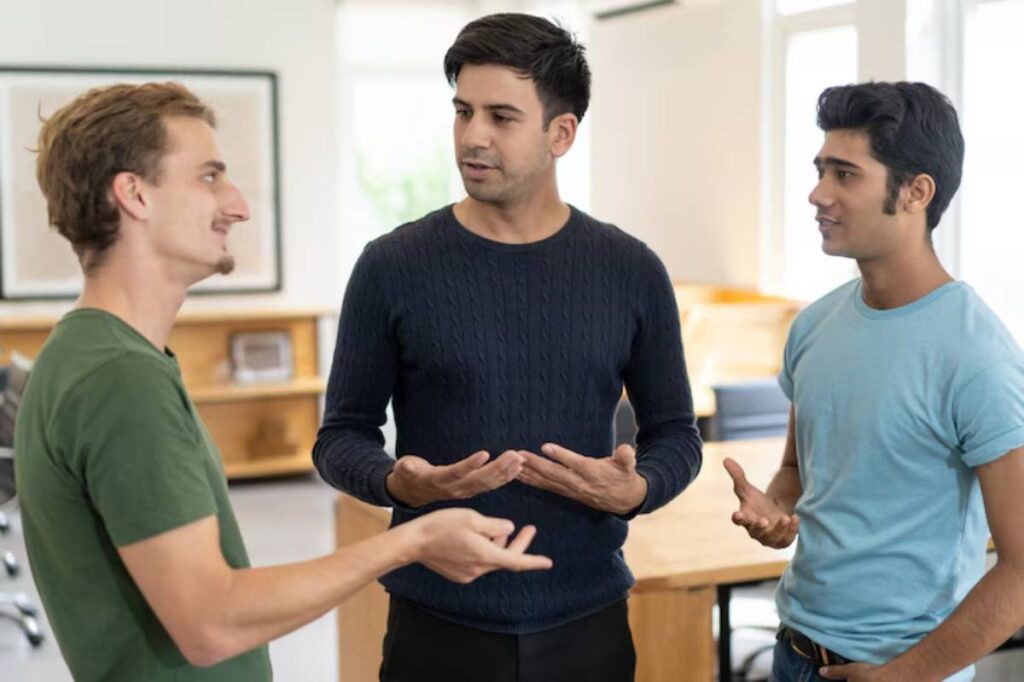 Three young men engage in a lively discussion with expressive hand gestures in a bright, modern office setting.