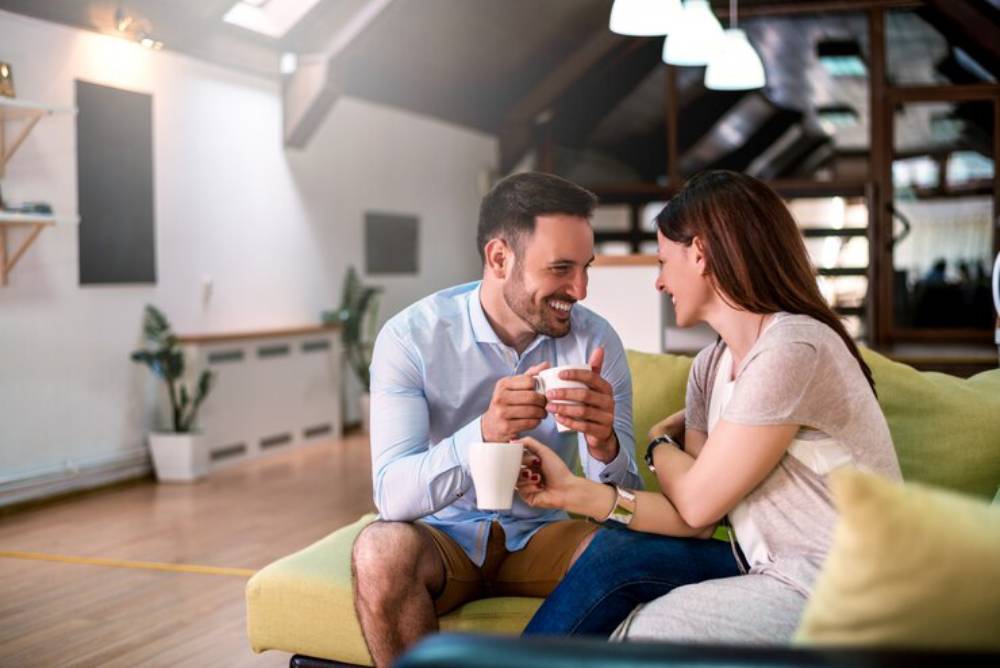 A young couple sharing an intimate moment on a green sofa in a cozy, well-lit living room, holding cups and smiling at each other.