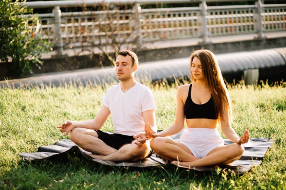 A couple meditating on yoga mats in a grassy area, surrounded by greenery and a soft, warm sunlight.