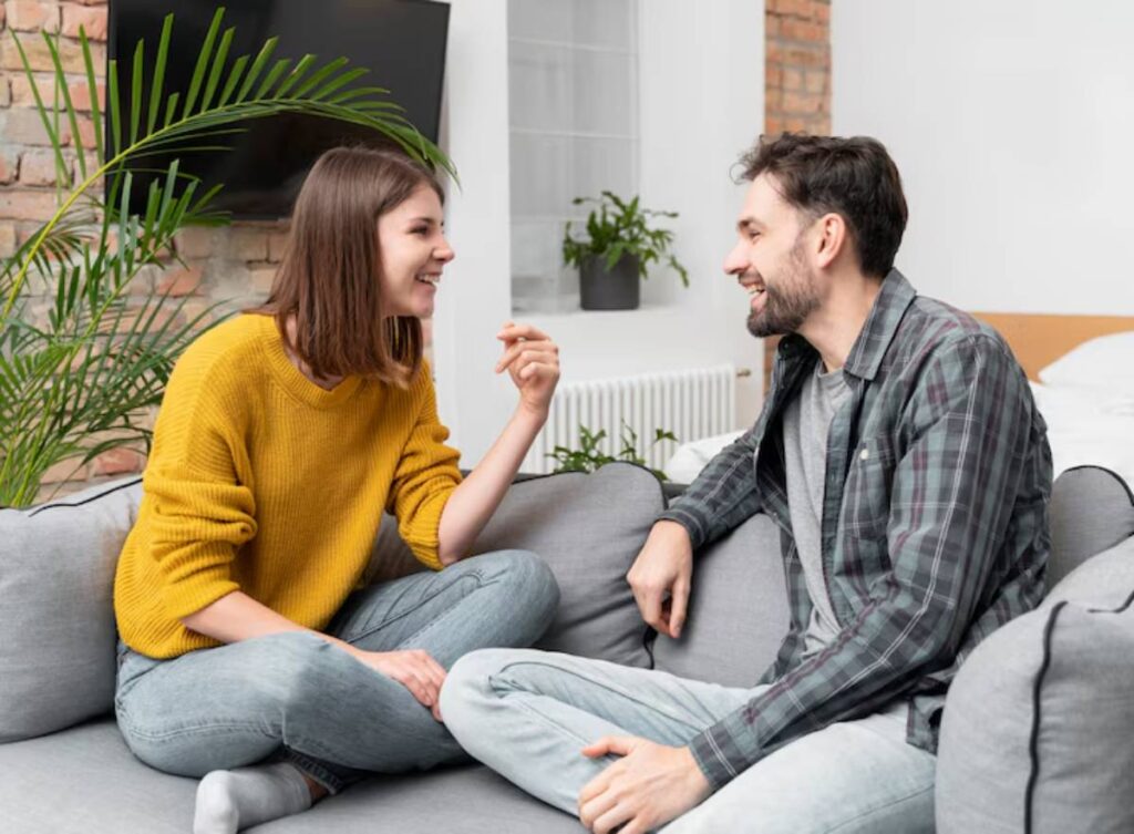 A couple sit on a gray couch, engaged in conversation in a modern, bright living room.